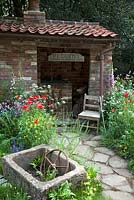 View of natural rock pathways and a brick shed and vintage chair surrounded by wildflowers. The Old Forge Artisan Garden for Motor Neurone Disease Association - RHS Chelsea Flower Show 2015