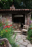 The Old Forge Artisan Garden for Motor Neurone Disease Association - RHS Chelsea Flower Show 2015. View of natural rock pathways and a brick shed and vintage chair surrounded by wildflowers