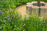 Thinking of Peace by Lace Landscapes. A circle of contemporary cylindrical stainless steel and wood seats under a tree surrounded by soft perennial planting of Iris sibirica 'Tropical Night' and 'Blue King', Veronica 'Pallida', Amsonia tabernaemontana, Luzula nivea, Geum 'Borisii'. 
