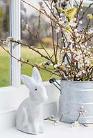 Floral display containing blossoming spring foliage in a galvanised bucket, a bunny and a view to the garden beyond