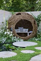 Spherical woven willow bird hide with bench, white flower borders with fencing, circular paving, chamomile lawn and pond with Nymphaea - Living Landscapes: City Twitchers Garden, RHS Hampton Court Palace Flower Show 2015