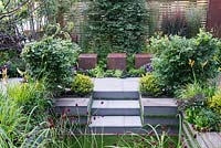 Metal cubed seating with steps down to sunken garden and artificial lawn. Pair of Fagus sylvatica and clipped Buxus sempervirens, Sanguisorba officinalis 'Morning Select' in foreground. Foundation for Growth Garden, RHS Hampton Court Palace Flower Show 2015