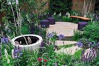 Stone pond in a pot water feature set in a flower bed, purple stools on a circular patio and a curved bench - The Wellbeing of Women Garden, RHS Hampton Court Palace Flower Show 2015 