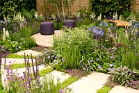 Curving path of stone slabs leading to circular seating area surrounded by flower beds planted with purple blue and white perennials and grasses. Agapanthus africanus Lupinus Salvia nemorosa Cardonna Chamaemelum nobile Pennisetum orientalis Digitalis Camelot White Salvia officinalis Purpurascens Thymus serphyllum Geranium Rozanne and Verbena rigida in The Wellbeing of Women Garden at RHS Hampton Court Palace Flower Show 2015 