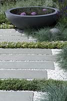 A large circular bowl of water with 3 Allium heads amongst slat and gravel paving interplanted with Thymes 'Snowdrift' and edged with Deschampsia 'Pixie Fountain'.  Designer: Rae Wilkinson, Sponsor Living Landscapes
