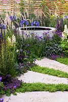 The Wellbeing of Women Garden, view of circular, concrete water feature and stepping stones path engraved with dates surrounded by Chamaemelum nobile, Thymus serpyllum, Agapanthus africanus, Verbena, white lupinus, Pennisetum orientalis, Salvia officinalis 'Purpurascens' against woodenn fence. Designers: Wendy von Buren, Claire Moreno, Amy Robertson. Sponsor: Tattersall Landscapes, London Stone, Jacksons Fencing, Hedgeworx, Tactile Studios