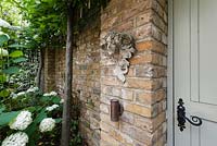 Walpole Gardens: London. July. Garden wall stone head sculpture. Carpinus betulus, Hydrangea arborescens 'Annabelle'