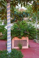 Painted tree in raised bed on red patio.  Tree painted in blue and white stripes. Alaior, Menorca. 
