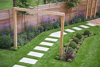 View of the path featuring white stone slabs, wooden beam entrance and borders with Verbena bonariensis, Buxus sempervirens, Delphinium elatum 'Blaustrahl' and Geranium 'Johnson's Blue'