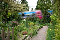 View from a garden gate down a gravel path beside rockery raised bed border with a train speeding by the bottom of the garden