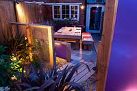 Contemporary long narrow small urban family garden with wooden decked paths at night. 