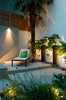 Small urban courtyard garden - outdoor living room with lounger under tall Chusan palm trachycarpus with wall lighters and plants in pots. 