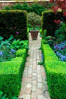 Brick path through low box hedge borders leading through a yew hedge towards focal point of standard holly in a container 