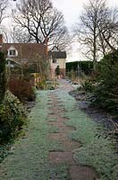 Frosty winter garden in February. Offset slab path through lawn between borders towards house 