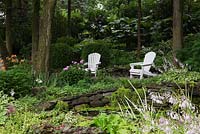 Two white wooden adirondack chairs on edge of pond with Nymphaea - Water Lily pads bordered by mauve flowering Hosta plants, Quebec, Canada.