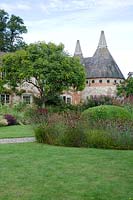 Bury Court with oast houses, perennial planting under a Koelreuteria paniculata tree, plants include Echinacea 'Fatal Attraction', Stipa tenuissima 'Pony Tails', Oreganum vulgare, clipped Buxus sempervirens surrounded by Dianthus carthusianorum