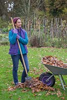 A woman leaning on a garden rake with a pile of raked autumnal leaves