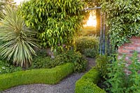 Early morning sun shines through a gateway between the Long Walk into the Croft Garden at Wollerton Old Hall Garden, Shropshire. The wrought iron gate is clothed in variegated ivies and a variegated Cordyline is planted close by. 