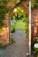 The dawn sun shines through a gate leading to The Yew Walk at Wollerton Old Hall Garden Shropshire. This area of the garden features tall, clipped yew pyramids together with roses, Salvia and Penstemons.