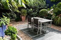 A garden room designed for entertaining with charcoal grey paving topped with a table and chairs, cobalt walls and bold leaves of palms and cordylines.
