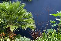 Chusan palm - Trachycarpus fortunei, Fatsia japonica and metal flower sculptures in front of a cobalt blue wall.