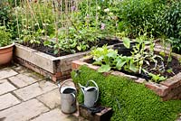 Raised beds for vegetables and for potting on young plants, made from red bricks and wooden sleepers with adjacent stone slab paving and tap with watering cans.   