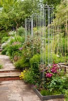 Border with iron obelisks for Clematis and Rosa in long mixed border with perennials shrubs and trees by stone path with steps in backyard garden in summer