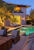 View of modern swimming pool and house with outside seating area sun loungers and mature olive tree at dusk.