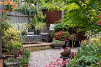 Tucked away in quiet corner at bottom of garden, between shed and greenhouse, a raised wooden deck with outdoor rattan furniture. Pots of acer, bonsai, heuchera and ornamental grasses. In tall pot Carex oshimensis 'Evergold' and berberis with nandina, mahonia and coleus.