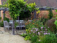 Dining table and chairs beneath pergola with wisteria and vine. To right, autumn bed of pineapple flower, Tithonia rotundifolia, Tulbaghia violacea and miscanthus.