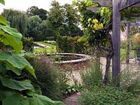 A view from the hot border with Wisteria, Vitis, Guara lindheimeri, Tithonia rotundifolia and Paulownia tomentosa to the raised brick pond and formal garden beyond.