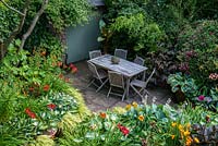A secluded shady patio with garden furniture surrounded by lush dense borders planted with Hostas, Hakonechloa grasses, Ligularia, Persicaria microcephala, Lilium martagon, daylilies, Clematis Etoile Violette, Bamboo and a banana plant 
