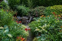 A small secluded seating area with garden furniture surrounded by lush dense borders planted with Hostas, Hackonechloa grasses, red daylilies, Lilium martagon with Clematis Etoile Violette, Bamboo and Persicaria microcephala at the far end.