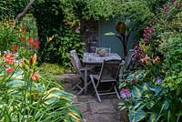 A small secluded seating area with garden furniture surrounded by lush dense borders planted with Hostas, Hakonechloa grasses, daylilies, Lilium martagon with Clematis Etoile Violette - right and a banana plant in a container at the far end.