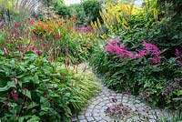 A small path leading through dense herbaceous borders including Hakonechloa macra Aureola, Persicaria Firetail and Calamagrostis - right with Astilbe Fanal and ligularia przewalskii - left. In the far border Crocosmia, Persicaria and Stipa gigantia.