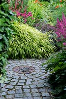 A stone set path in a circular design lined with dense herbaceous planting including yellow Hakonechloa macra Aureola grass, Persicaria Firetail, Monarda, Sedum, black Ophiopogon and Astilbe.