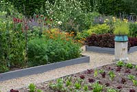 A potager with raised beds of vegetables and flowers including chives, salad leaves, peas, marigolds, bergamot and daylilies.A stone plith with a copper pot of lavender provides a focal point.