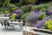 A patio seating area, water feature and large bank planted with perennials and ornamental grasses. Planting includes: Salvia verticaillata 'Purple rain', Geranium 'Rozanne', Campanula persicifolia, Salvia 'Caradonna' and Molinia 'Karl Foerster'