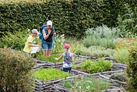 Mother and children in the formal herb garden, with planted squares woven around the sides and sheltered by hedges at Jardin des Cimes, Chamonix, France. July 