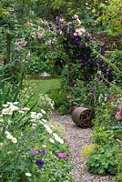 A country cottage garden with Rosa 'Belvedere', Rosa 'Compassion' and Clematis 'Etoile Violette' scrambling over a rustic arch which frames view of potager beyond. Pebble path edged in borders of leucanthemum, alchemilla, hardy geranium, fuchsia and Jacob's ladder.