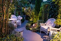 A small contemporary garden at night, with curved wooden path and seating area lit at night.