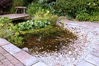 Rectangular wildllife pond adjacent to raised patio and edged with stone and brick paving and with shallow end with pebbles and cobbles  