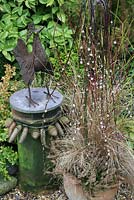 Muted brown and bronze tones for winter and spring. Rust coated cockerel sculpture made from scrap metal and showcased on a chimney pot topped with a circle of slate and decorated with a necklace of fir cones. Weeping pussy willow is underplanted with bud heathers and brown leaved sedge in a terracotta pot.
