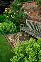 A walled backed garden seat set on a pebble plinth and flanked by Lady's Mantle, Alchemilla mollis and standard Rosa Graham Thomas. June. Worcestershire