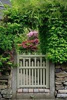 A mixed country - style hedge of hornbeam, Lonicera nitida, hawthorn and purple leaved Berberis has been trained to form an arch over a gate that frames a clump of Azaleas on the path to the front door.