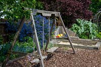 A wooden swing and sand pit made from railway sleepers in a children's play area with floor covered with bark chippings. Behind, grey leaved cardoon and blue ceanothus.
