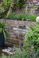 Water spills out from the side of a brick wall, splashing onto the pebbles below. Raised bed planted with wallflower, lavender, pink cosmos, salvia and black grass.