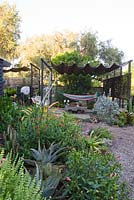 View into seating area of garden showing chairs and hammock. In the foreground: red flowers of Ruellia elegans, behind it yellow foliage of Aechmea blanchetiana, Ocimum basilicum - basil flowers, agave and native hibiscus
