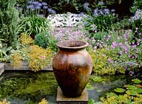 Small formal pond with empty urn ornament in centre, wooden edging, nymphaea, alchemilla mollis, geranium, agapanthus, August. Lexley Duncan's garden, London