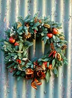 Close up of blue pine wreath with chillies, crab apples, dried fruit, cones, cinnamon sticks, seasonal berries, lotus heads eucalyptus pods, sisal and ribbon bows on corrugated iron background. Design: Elaine Boon at Pesh Flowers.